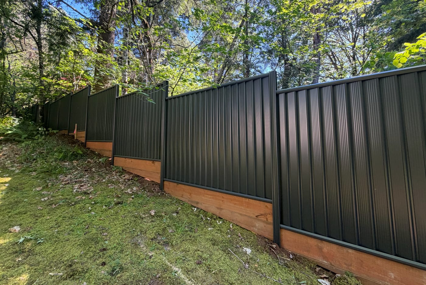 Dark green Colorbond steel fence stepping down a sloped, wooded yard in the Portland, OR area, built by Gorilla Works on a cedar kickboard